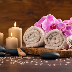 Spa setting with brown rolled towel, orchids and candles on wood. Relaxing spa concept with candles, towels and hot stones massage with himalayan pink salt. Beautiful composition for beauty treatment in a spa.